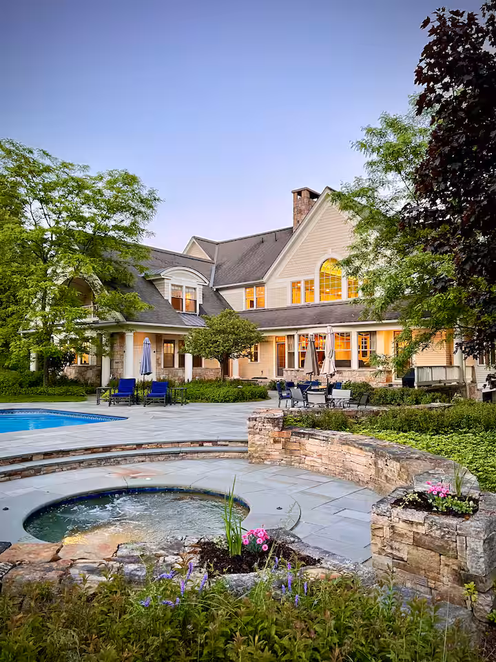Rear exterior twilight view of The Lemelson Estate, showcasing the glowing warm windows overlooking the stone-paved patio and private swimming pool.